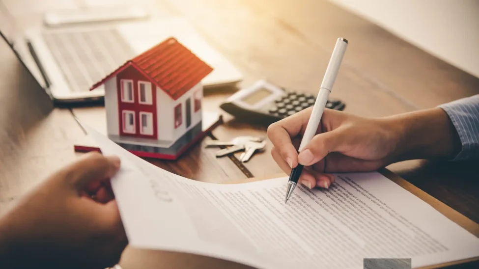 A person signing a contract related to types of construction bonds, with a model house, calculator, and keys on a wooden desk.