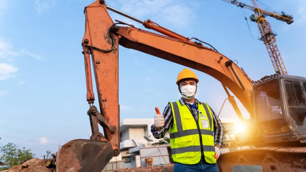 A construction worker wearing a safety vest and helmet gives a thumbs up while standing in front of an orange excavator, one of the types of construction equipment, at a construction site.