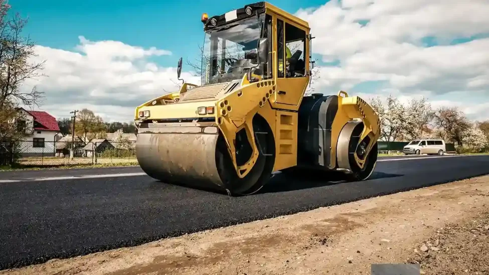 Under a partially cloudy sky, a yellow road roller compacts recently put asphalt on a rural road, exhibiting types of road construction techniques amid homes and blossoming trees.