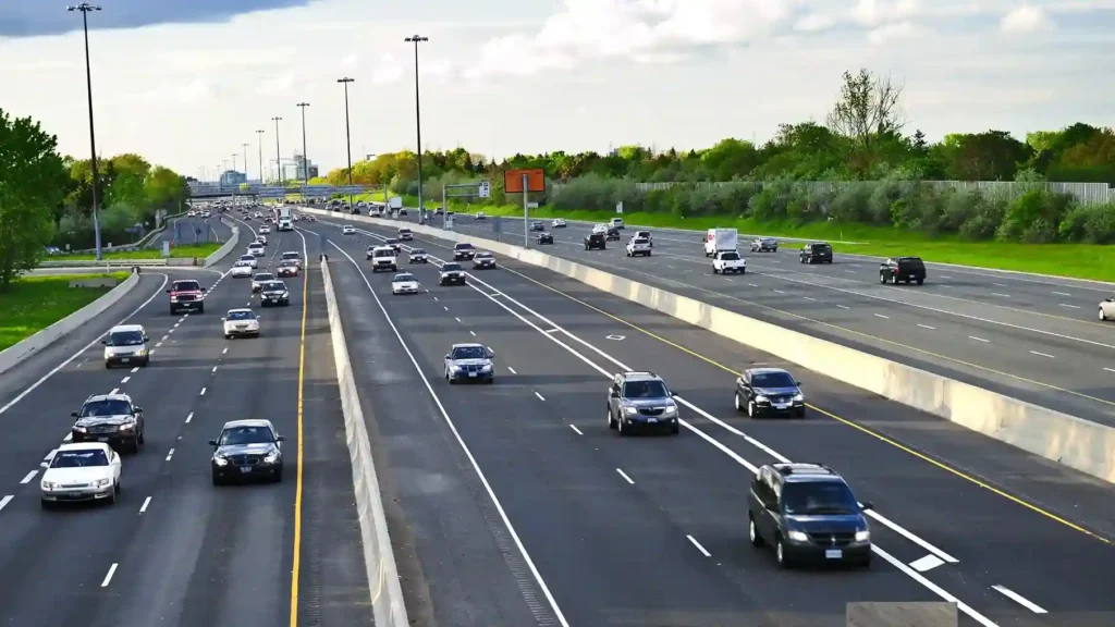 A multi-lane highway with flowing traffic, surrounded by greenery and urban structures under a bright, partly cloudy sky, representing the outcomes of modern types of road construction.