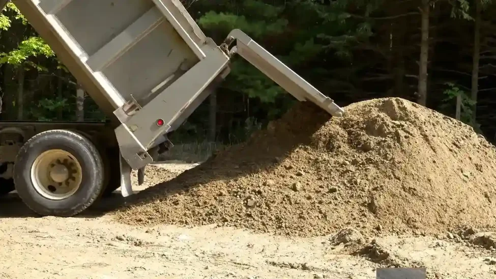 A truck unloading a large pile of sand used in construction at a site surrounded by trees.